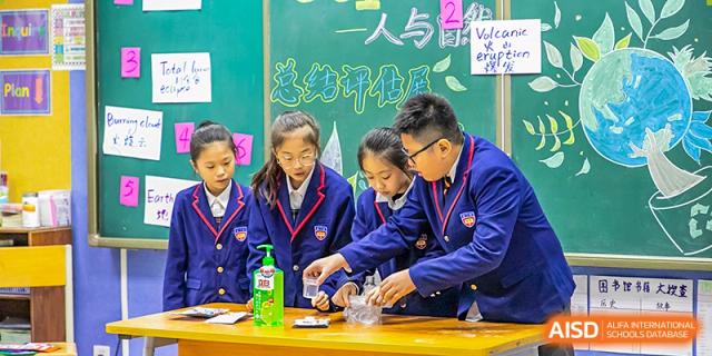 A group of older students in their school uniforms collaborating on a science experiment at a table with a chalkboard in the background at Wuhan Aoxin Elite School.