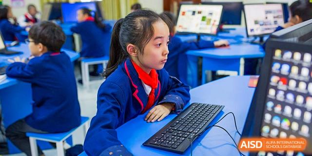 A young female student in a blue school uniform and red scarf focused on working with a keyboard and computer in a class at Wuhan Aoxin Elite School.