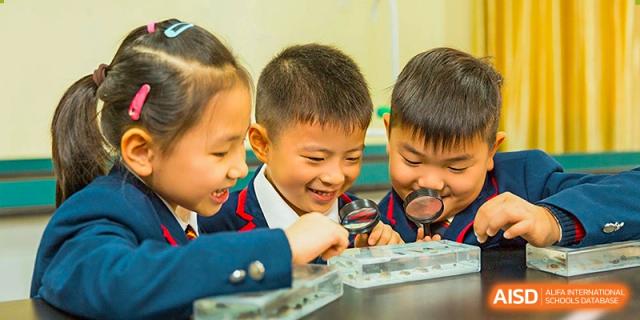 A group of young students smiling as they observe something closely using a magnifying glass during a hands-on learning activity at Wuhan Aoxin Elite School.