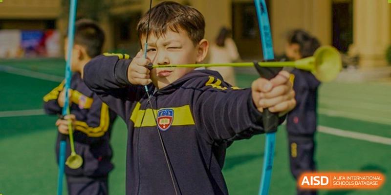 A young male student in a dark blue and yellow uniform aiming an archery bow and arrow during a sports activity at Wuhan Aoxin Elite School.