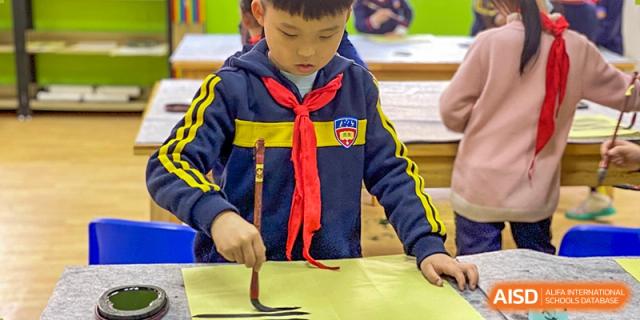 A young male student in a dark blue uniform and red scarf practicing Chinese calligraphy with a brush on yellow paper at Wuhan Aoxin Elite School.