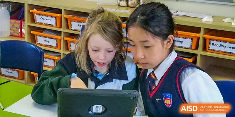 Two female students, one Western and one Asian, in school uniforms looking intently at a tablet or laptop screen together in a bright area at Wuhan Aoxin Elite School.