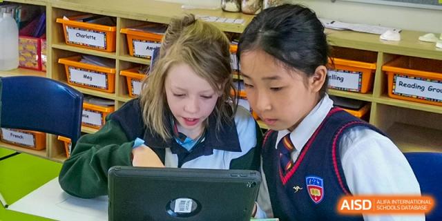 Two female students, one Western and one Asian, in school uniforms looking intently at a tablet or laptop screen together in a bright area at Wuhan Aoxin Elite School.