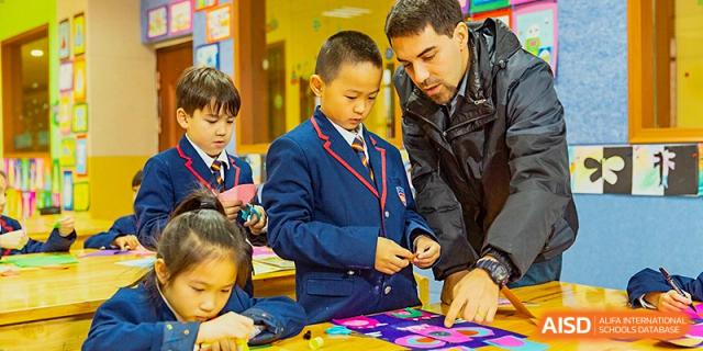 An adult male teacher kneeling to assist a young male student in a blue blazer with a hands-on activity, possibly involving crafts or design, at Wuhan Aoxin Elite School.