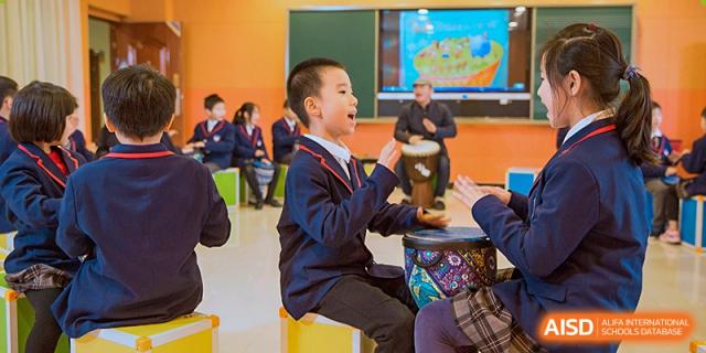 A young student in a school uniform laughing and playing a small hand drum during a music class or activity at Wuhan Aoxin Elite School.
