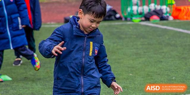 A young boy in a blue jacket looking down and interacting with something on a green field during outdoor play at Wuhan Aoxin Elite School.