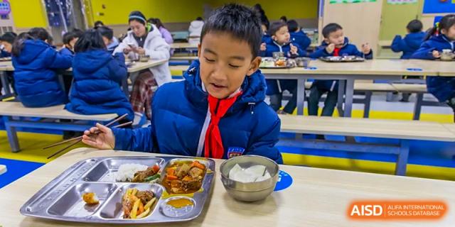 A male student in a blue jacket and red scarf happily eating a meal from a segmented tray and a metal bowl in the school cafeteria at Wuhan Aoxin Elite School.