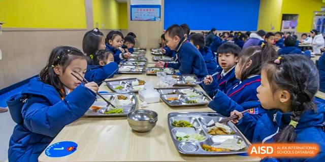 A wide shot of several students in uniform seated at tables, eating lunch in the school cafeteria at Wuhan Aoxin Elite School.