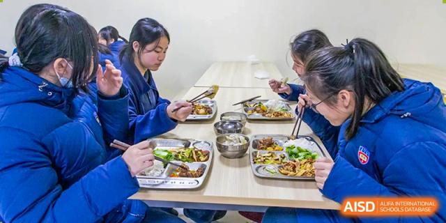 Two female students seated at a table, eating their separate meals from segmented trays and bowls, suggesting a shared mealtime experience at Wuhan Aoxin Elite School.
