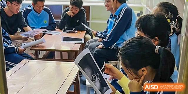 Students from the Wuhan Hongshan High School International Department in class, one holding up a large printout and interacting with classmates.