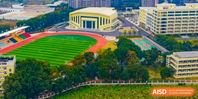 Wuhan Hongshan High School International Department - Aerial view showing a yellow building next to a red running track and green sports field, surrounded by trees and foliage.