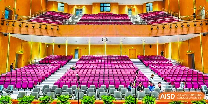 Vibrant auditorium interior. Wuhan Hongshan High School International Department. Rows of bright pink seats, stage area, yellow accents. Modern hall for events/gatherings.
