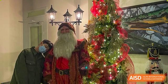 Santa Claus stands by a Christmas tree. Wuhan Hongshan High School International Department. Close-up of a person dressed as Santa, lights and decorations, holiday celebration.