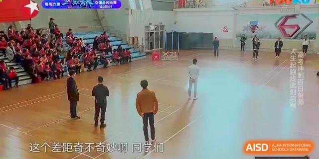 Two students in an indoor gym. Wuhan Hongshan High School International Department. A young man in a yellow jacket walking on a wooden floor court, with a group in the background.