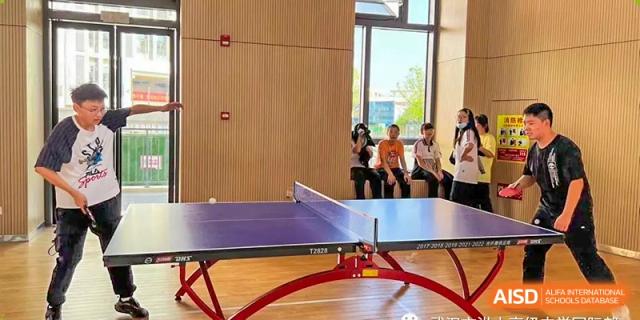 A table tennis (ping pong) table setup. Wuhan Hongshan High School International Department. Students are sitting nearby in a brightly lit room with wooden panel walls and large windows.