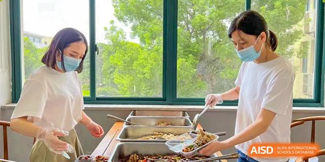 A close-up of a person serving food from a buffet line with trays of prepared dishes at Wuhan Hongshan High School International Department.