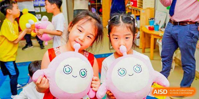 Two young female students from BASIS International School Guangzhou smiling and holding pink, round, smiling plush toys.