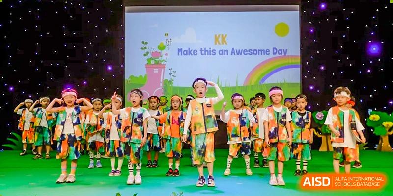 A group of young students from BASIS International School Guangzhou in colorful outfits performing on a stage with a banner that reads "KK Make this an Awesome."