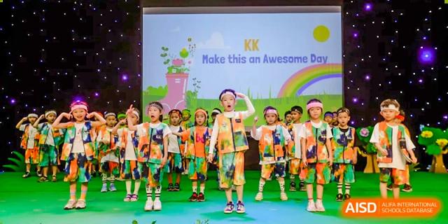 A group of young students from BASIS International School Guangzhou in colorful outfits performing on a stage with a banner that reads "KK Make this an Awesome."