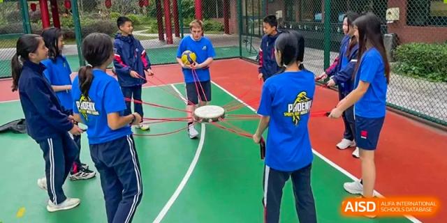 Students at BASIS International School Guangzhou participating in an outdoor team-building game with a disc and ropes on a green and red sports court.