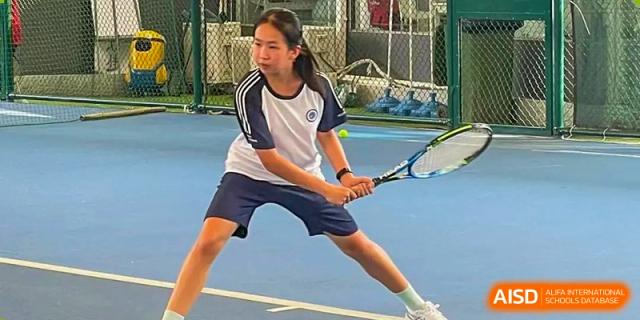 A female tennis player from BASIS International School Guangzhou in a blue and white uniform poised to return a shot.