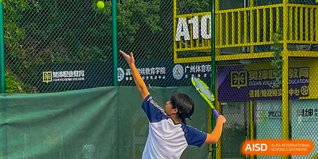 A female student from BASIS International School Guangzhou in a blue and white uniform winding up to hit a tennis serve on an outdoor court.
