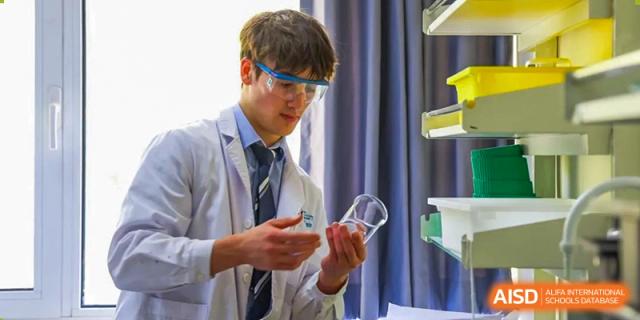 A male student from Harrow International School Beijing (AISL Beijing) wears a lab coat and safety goggles while examining laboratory equipment.