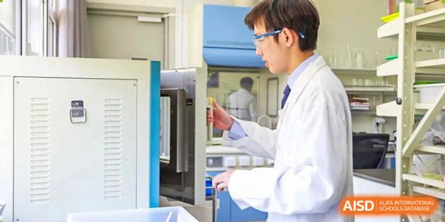 A male student from Harrow International School Beijing (AISL Beijing), dressed in a lab coat and goggles, is working with lab equipment.