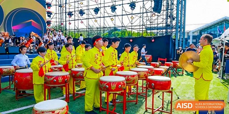 A group of students from Harrow International School Beijing (AISL Beijing) are performing a traditional drum dance outdoors, dressed in vibrant yellow costumes.