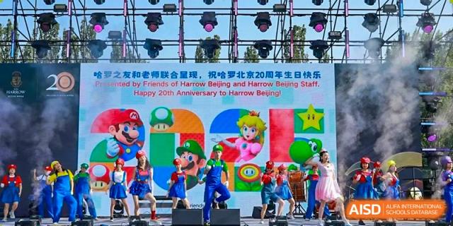 Students from Harrow International School Beijing (AISL Beijing) are performing on a brightly lit stage with colorful backdrops featuring Mario characters, likely for an anniversary celebration or school show.