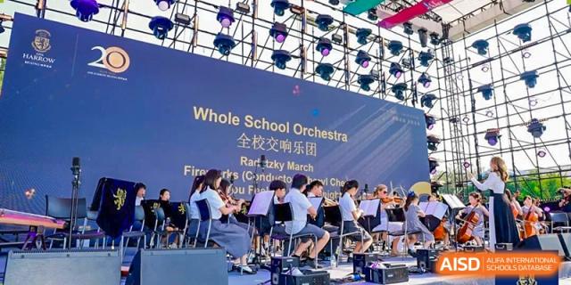 The Harrow International School Beijing (AISL Beijing) Whole School Orchestra is performing on an outdoor stage with professional lighting.