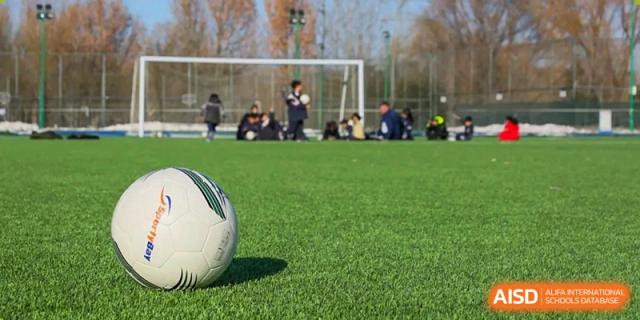 A football rests on a green sports field at Harrow International School Beijing (AISL Beijing), with students visible in the background.