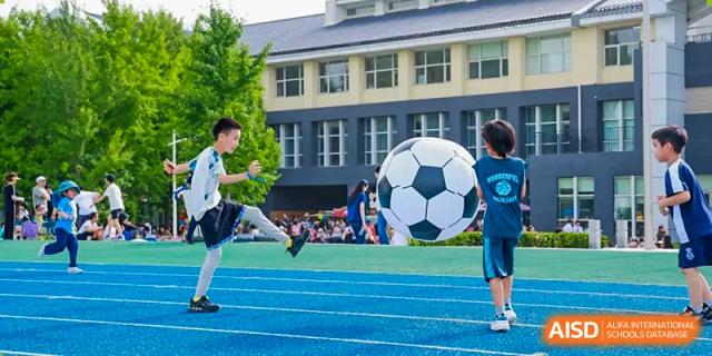 Two students from Harrow International School Beijing (AISL Beijing) are playing on a sports field with a large, oversized soccer ball.