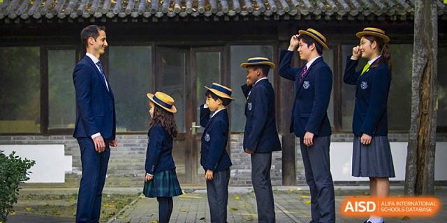 A group of students from Harrow International School Beijing (AISL Beijing), dressed in their school uniforms, are standing in a line and saluting an adult.