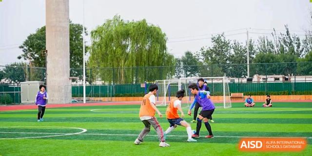Students in orange and purple jerseys playing soccer on a green turf field with a large willow tree in the background at BEIJING CHAOYANG TONGWEN FOREIGN LANGUAGE SCHOOL.