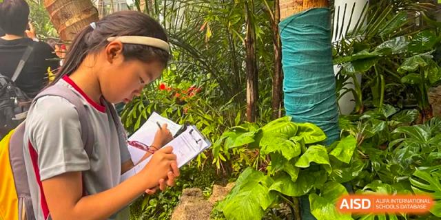 A student taking notes outdoors while surrounded by lush tropical plants and green leaves. Beanstalk International Bilingual School (BIBS) - Chengdu