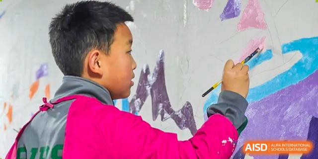 A close-up of a student in a pink jacket focused on painting a large mural with a brush. Beanstalk International Bilingual School (BIBS) - Chengdu