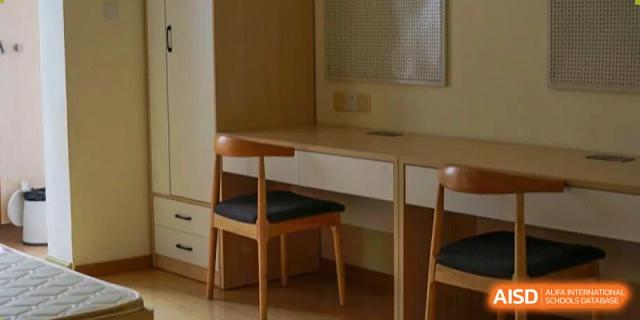 A minimalist student dormitory featuring a light wood study desk, modern wooden chairs with black cushions, and built-in white storage drawers at Beanstalk International Bilingual School (BIBS) - Chengdu.