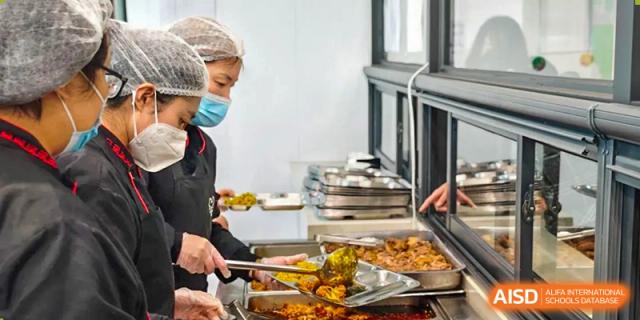 Staff members wearing masks and hairnets serve nutritious hot meals into metal trays for students in a clean, professional cafeteria at Beanstalk International Bilingual School (BIBS) - Chengdu.