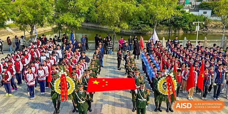 A grand procession of military personnel carrying the Chinese national flag past a crowd of onlookers at an outdoor event for The Experimental School Affiliated with Zhuhai No.1 High School.
