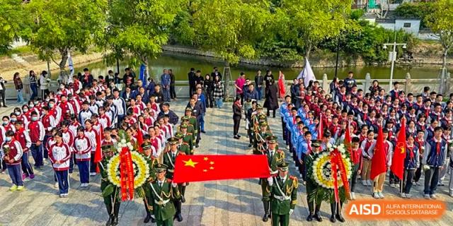 A grand procession of military personnel carrying the Chinese national flag past a crowd of onlookers at an outdoor event for The Experimental School Affiliated with Zhuhai No.1 High School.
