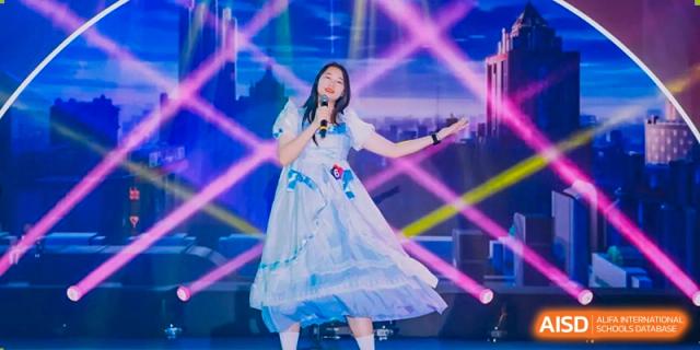 A student from The Experimental School Affiliated with Zhuhai No.1 High School performing a song on stage in a flowing light blue and white dress.