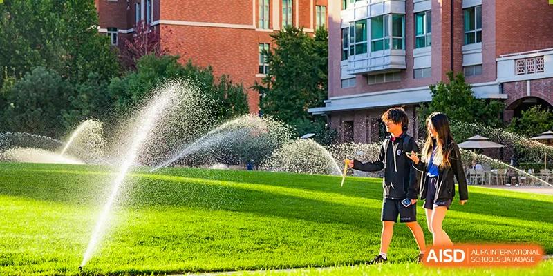 A Beijing Keystone Academy student in a uniform playing in the spray of a sprinkler on a sunny day.
