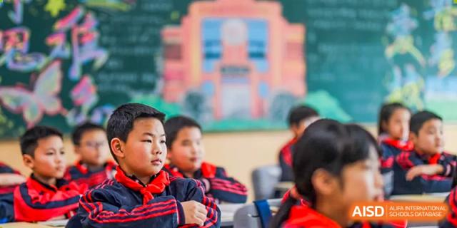 A close-up of young students from Changsha Lushan Zhongjia School in red tops, looking ahead during an activity, with the main school building blurred in the background.