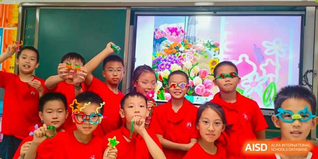 A group of young students from Changsha Lushan Zhongjia School in red shirts celebrating an activity or presentation with flowers and props in an excited group huddle.
