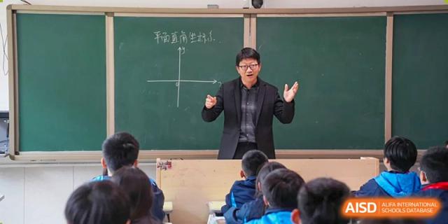 A male teacher from Changsha Lushan Zhongjia School wearing glasses and a suit, enthusiastically teaching a lesson at a green chalkboard with a graph drawn on it.