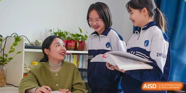 A female teacher and a female student from Changsha Lushan Zhongjia School smiling and engaging in a discussion over a book in a library or classroom setting.