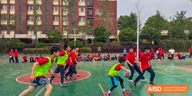 Students from Changsha Lushan Zhongjia School participating in an outdoor sports game on a green court, with a large, multi-story brick school building visible behind them.