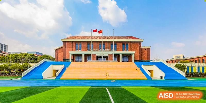 The grand, traditional main teaching building of Changsha Lushan Zhongjia School, featuring red brick architecture and a spacious entrance, overlooking the blue and green athletic field.