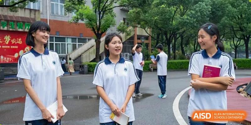 A female student from Changsha Lushan Zhongjia School wearing a white school uniform, smiling and holding papers while standing outdoors on a paved area with other students in the distance.